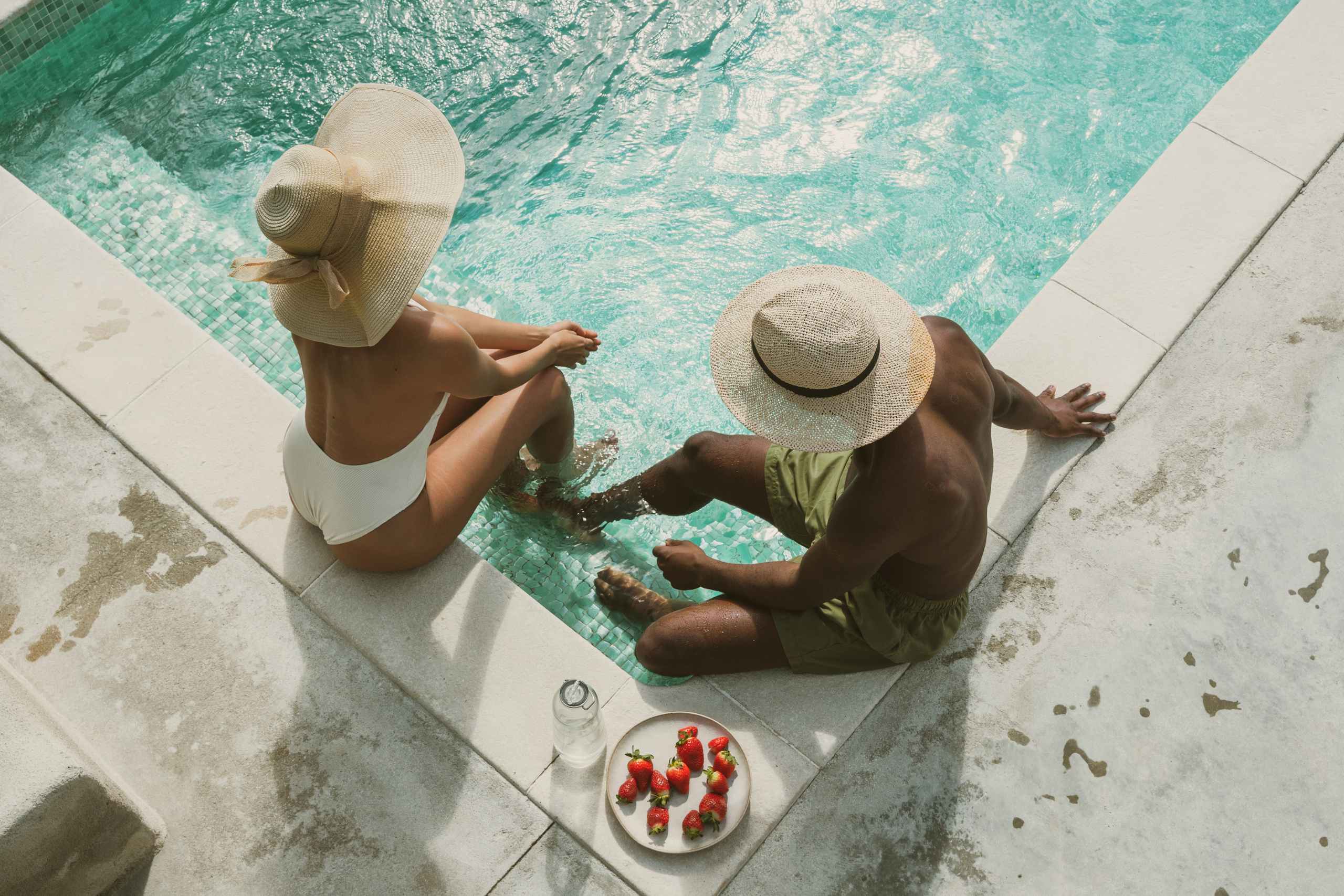 Two people in swimsuits sit at the edge of a turquoise pool, wearing wide-brimmed hats, with a plate of strawberries and a water bottle nearby on the pool deck.